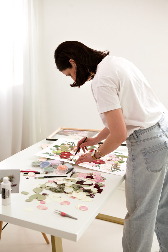 Mitarbeiterin arrangiert gepresste Blüten für ein Brautstrauß-Erinnerungsbild. Sie arbeitet an einem weißen Tisch mit gepressten Blumenblättern in Rosa, Rot und Grün. Verschiedene Farbtöpfchen zum Nachfärben der Blüten stehen bereit. Professionelle Brautstraußkonservierung in Leipzig.