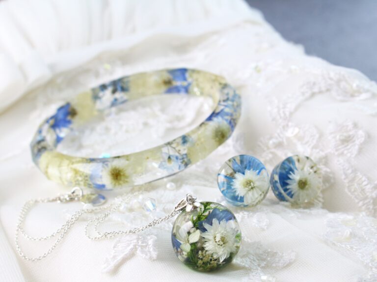 Eleganter Schmuckset aus Epoxidharz mit eingebetteten blauen und weißen Blumen: Armreif, Anhänger an Silberkette und Ohrringe, platziert auf weißer Spitze