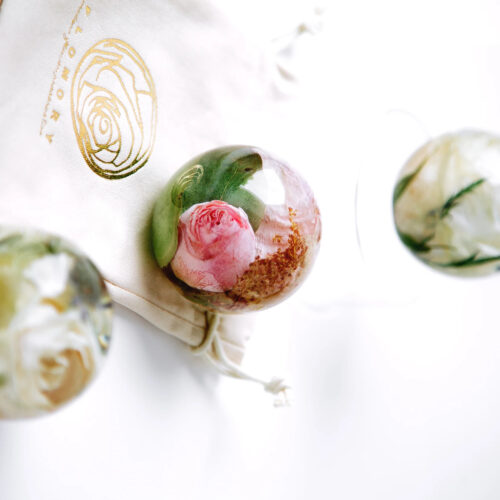 Drei transparente Kunstharz-Kugeln mit konservierten Blumen im Inneren, arrangiert auf einem cremefarbenen Stofftuch mit goldenem Logo. Die mittlere Kugel enthält eine rosa Rose mit grünen Blättern, während die obere und untere Kugel weiße Blüten mit grünem Blattwerk zeigen. Diese dekorativen Objekte dienen als Erinnerungsstücke, um besondere Blumen, möglicherweise aus einem Brautstrauß, dauerhaft zu bewahren. Die sphärische Form ermöglicht eine vollständige Betrachtung der konservierten Blüten von allen Seiten und schafft ein elegantes, zeitloses Andenken.