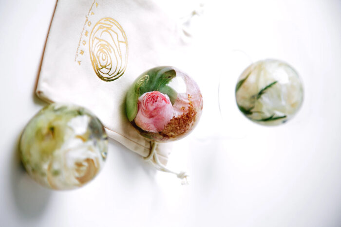 Drei transparente Kunstharz-Kugeln mit konservierten Blumen im Inneren, arrangiert auf einem cremefarbenen Stofftuch mit goldenem Logo. Die mittlere Kugel enthält eine rosa Rose mit grünen Blättern, während die obere und untere Kugel weiße Blüten mit grünem Blattwerk zeigen. Diese dekorativen Objekte dienen als Erinnerungsstücke, um besondere Blumen, möglicherweise aus einem Brautstrauß, dauerhaft zu bewahren. Die sphärische Form ermöglicht eine vollständige Betrachtung der konservierten Blüten von allen Seiten und schafft ein elegantes, zeitloses Andenken.