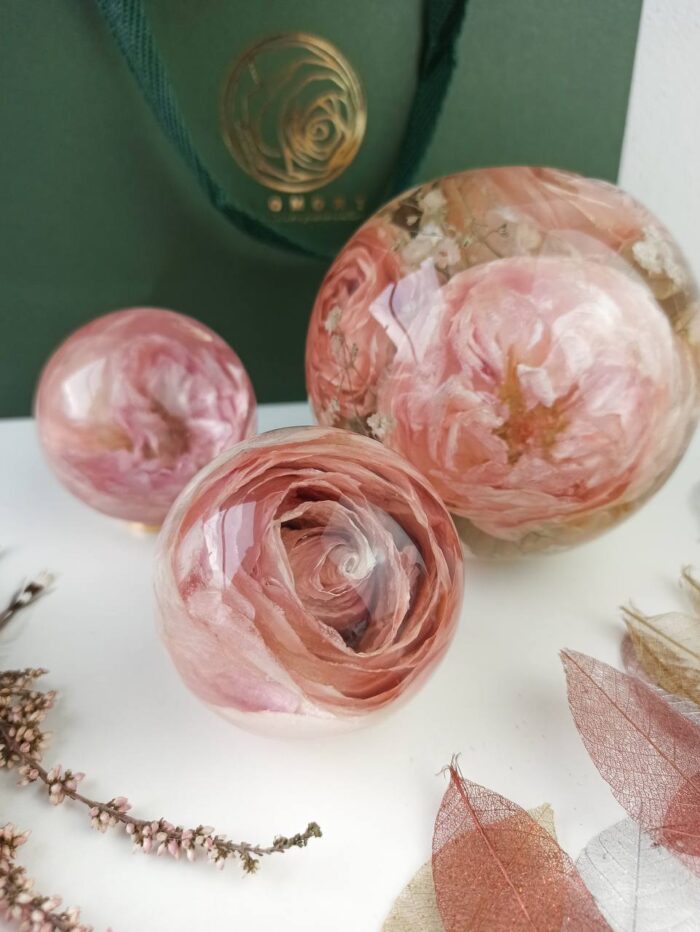 Drei transparente Kunstharzkugeln mit konservierten rosafarbenen Rosen in verschiedenen Größen, arrangiert auf einer weißen Oberfläche. Die Kugeln zeigen detaillierte Blütenstrukturen in zarten Rosa- und Korallentönen, perfekt in klarem Harz konserviert. Im Hintergrund ist eine dunkelgrüne Tasche mit einem goldenen Rosen-Logo zu sehen. Die Komposition wird durch dekorative Elemente wie filigrane Skelettblätter und getrocknete Pflanzenzweige am unteren Bildrand ergänzt. Diese Kunstharzkreationen dienen als dauerhafte Erinnerungsstücke und dekorative Objekte, die die natürliche Schönheit von Blumen bewahren, häufig aus Brautsträußen oder bedeutsamen Anlässen.