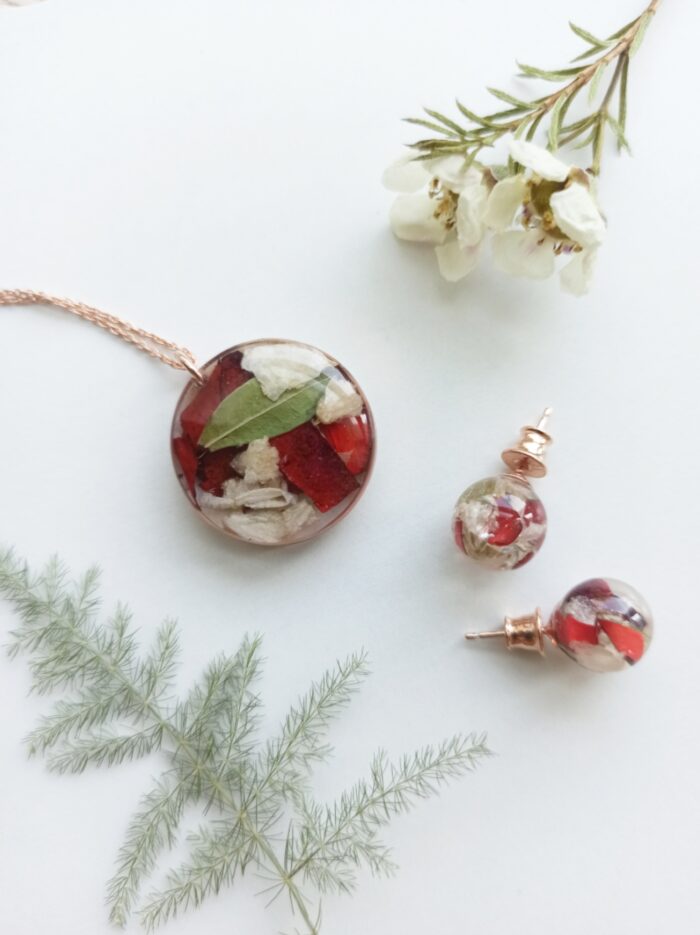Ein elegantes Schmuckset aus Brautstraußblumen in Kunstharz mit roségoldenen Elementen auf weißem Hintergrund. Das Set besteht aus einer runden Halskette mit geflochtener Roségoldkette und passenden Ohrsteckern. In den transparenten Kunstharzkugeln sind konservierte Blütenelemente in Rot, Weiß und Grün eingebettet – vermutlich Teile eines Brautstraußes mit roten Rosenblütenblättern, weißen Blüten und grünen Blättern. Die Komposition wird durch frische Grünelemente und kleine weiße Blüten im oberen und unteren Bildbereich umrahmt. Diese Art von personalisiertem Schmuck ermöglicht es Bräuten, Teile ihres Hochzeitsstraußes als dauerhafte Erinnerung zu bewahren und zu tragen.