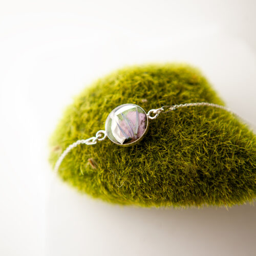 Silbernes Armband Barcelona mit rundem Resin-Anhänger, der gefriergetrocknete Brautblumen in Rosa und Grün einschließt, drapiert auf einem moosbedeckten Stein
