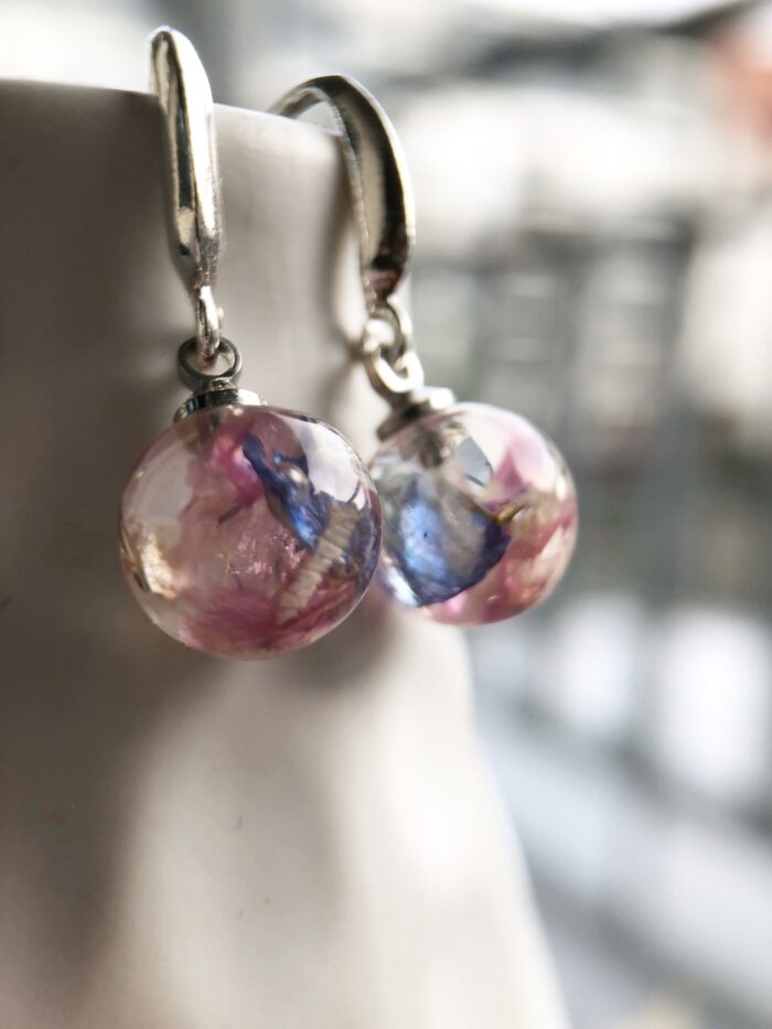 Ein Paar elegante Ohrringe mit transparenten Kunstharzkugeln an silbernen Bügeln. In den runden Kugeln sind zarte rosa und blaue getrocknete Blütenstücke in klarem Harz konserviert, die ein verträumtes, mehrfarbiges Erscheinungsbild erzeugen. Die Ohrringe hängen an einer hellen Oberfläche vor einem unscharfen Hintergrund. Die handgefertigten Schmuckstücke kombinieren die natürliche Schönheit der eingebetteten Blütenelemente mit modernem Design und eignen sich perfekt für Alltags- oder festliche Anlässe.