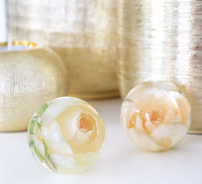 Zwei transparente Kunstharzkugeln mit konservierten cremefarbenen Rosen im Inneren, auf weißer Oberfläche platziert. Die linke Kugel zeigt eine vollständig sichtbare cremefarbene Rose mit zarten grünen Blättern, während die rechte Kugel eine ähnliche, leicht verschwommene Rose enthält. Im Hintergrund sind weiche, goldene Stofftexturen zu sehen. Diese dekorativen Objekte dienen der dauerhaften Konservierung von Blüten als elegante Erinnerungsstücke und werden häufig für die Aufbewahrung besonderer Andenken wie Hochzeitsblumen verwendet.