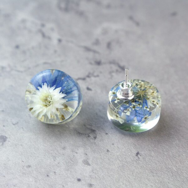 Handgefertigte Ohrstecker aus Epoxidharz mit weißer Blüte und blauen Akzenten, in Nahaufnahme auf grauem Hintergrund, zeigt silberne Verschlüsse und Details der eingebetteten Blumen