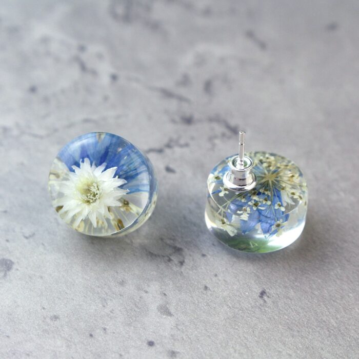 Handgefertigte Ohrstecker aus Epoxidharz mit weißer Blüte und blauen Akzenten, in Nahaufnahme auf grauem Hintergrund, zeigt silberne Verschlüsse und Details der eingebetteten Blumen