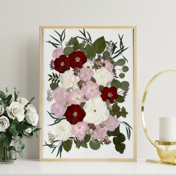 Großes gerahmtes Erinnerungsbild mit kunstvoll arrangierten gepressten Blüten aus einem Brautstrauß in Bordeauxrot, Rosa und Weiß mit grünen Blättern und Eukalyptus, präsentiert in einem schlanken schwarzen Rahmen auf einem modernen schwarzen Sideboard mit gerippter Front. Im stilvollen Wohnambiente mit weißer Tischlampe, dekorativer Muschelskulptur und Zimmerpflanzen in schlichten Keramiktöpfen.