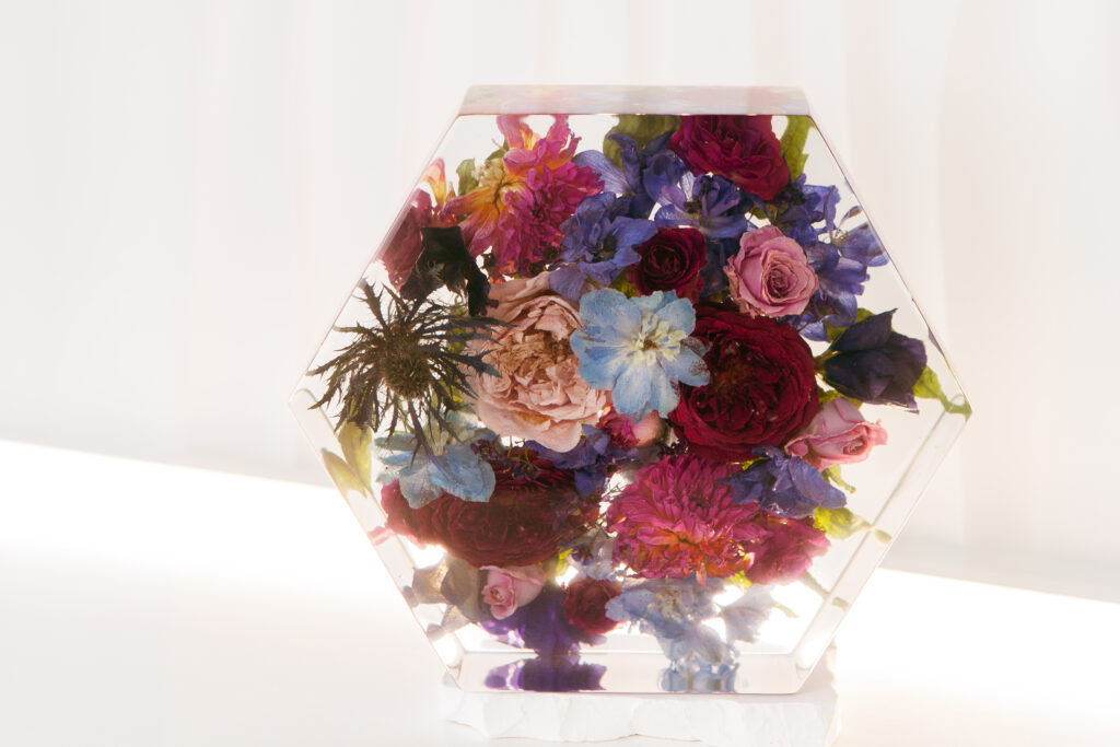 Gefriergetrockneter Brautstrauß in hexagonalem Resin-Block mit bordeauxroten Rosen, blauen Rittersporn-Blüten, pinken Pfingstrosen und Disteln in kristallklarem Harz