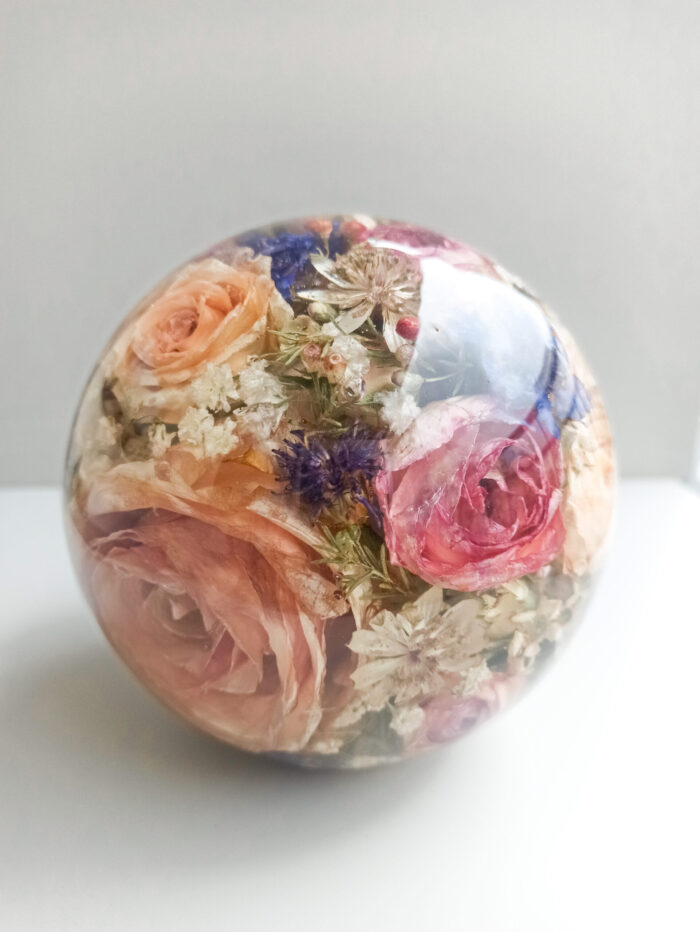 Eine transparente Kunstharzkugel mit einem farbenreichen Arrangement getrockneter Blumen auf weißer Oberfläche vor hellgrauem Hintergrund. Die Kugel enthält konservierte Rosen in verschiedenen Pastelltönen – apricot, rosa und pfirsich – zusammen mit lila Kornblumen und feinen weißen Schleierkrautblüten. Die Blumen sind in unterschiedlichen Ebenen im klaren Harz eingebettet, was einen dreidimensionalen Effekt erzeugt. Diese Konservierungsmethode bewahrt die natürliche Schönheit eines Blumenstraußes, wie etwa eines Brautstraußes, und transformiert ihn in ein dauerhaftes Erinnerungsstück. Die sanften Vintage-Farbtöne der getrockneten Blüten verleihen dem Objekt eine romantische, zeitlose Ästhetik.