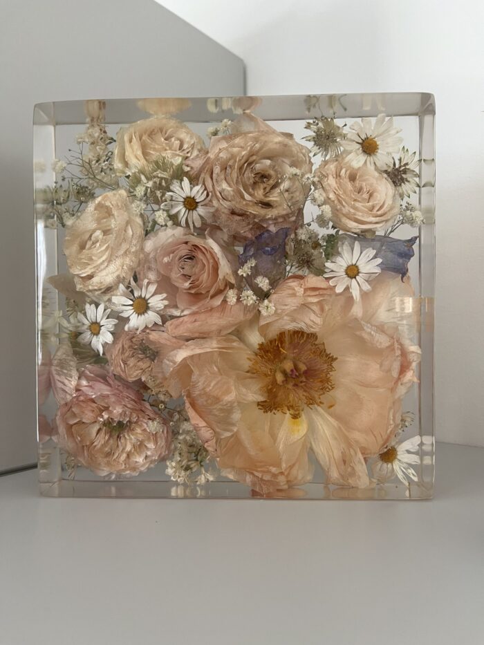 Ein quadratischer transparenter Kunstharzblock mit einem sorgfältig konservierten Brautstrauß in zarten Vintage-Pastelltönen. Der Block enthält mehrere pudrige apricotfarbene und blassrosa Rosen, eine große Pfingstrose mit goldener Mitte, weiße Gänseblümchen mit gelben Zentren, Schleierkraut und feine bläuliche Akzente. Die getrockneten Blumen sind präzise in kristallklarem Kunstharz eingebettet, was ihre natürliche Form und die zarte Farbgebung perfekt bewahrt. Der Block steht auf einer weißen Oberfläche vor einer neutralen grauen und weißen Wand. Diese professionelle Brautstraußkonservierung bietet eine dauerhafte Erinnerung an den Hochzeitstag und verwandelt den vergänglichen Blumenstrauß in ein zeitloses Dekorationsobjekt mit romantischer, nostalgischer Ausstrahlung. Die sorgfältige Trocknung und Einbettung erhält die charakteristische Textur und die subtilen Farbnuancen jeder einzelnen Blüte des Brautstraußes.