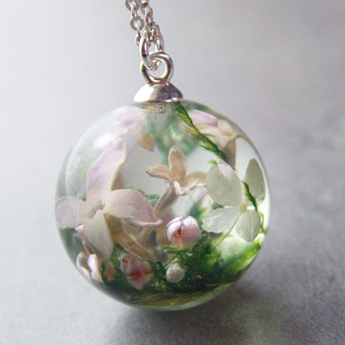 Ein runder Kunstharzanhänger an einer silbernen Kette, der wie ein winziges Terrarium gestaltet ist und Blumen aus einem Brautstrauß enthält. In der transparenten Kugel sind zarte rosa und weiße Blüten, kleine Knospen und grüne Moosstrukturen zu sehen, die eine natürliche Miniaturlandschaft bilden. Der Anhänger ist mit einer silbernen Öse befestigt und liegt auf einer grauen, strukturierten Oberfläche. Die verschiedenen Schichten der konservierten Pflanzenelemente erzeugen eine dreidimensionale Tiefenwirkung im klaren Harz. Diese Art von handgefertigtem Schmuck bietet eine kreative Möglichkeit, die Blumen des Hochzeitsstraußes als dauerhaftes Erinnerungsstück zu bewahren.RetryClaude can make mistakes. Please double-check responses.