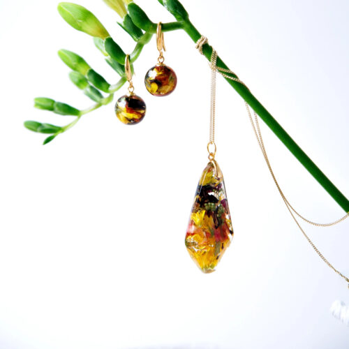 Goldenes Schmuckset mit facettiertem diamantförmigem Anhänger und passenden runden Ohrringen, alle mit bunten konservierten Blüten in Kunstharz, elegant präsentiert an einem grünen Pflanzenstängel vor weißem Hintergrund.