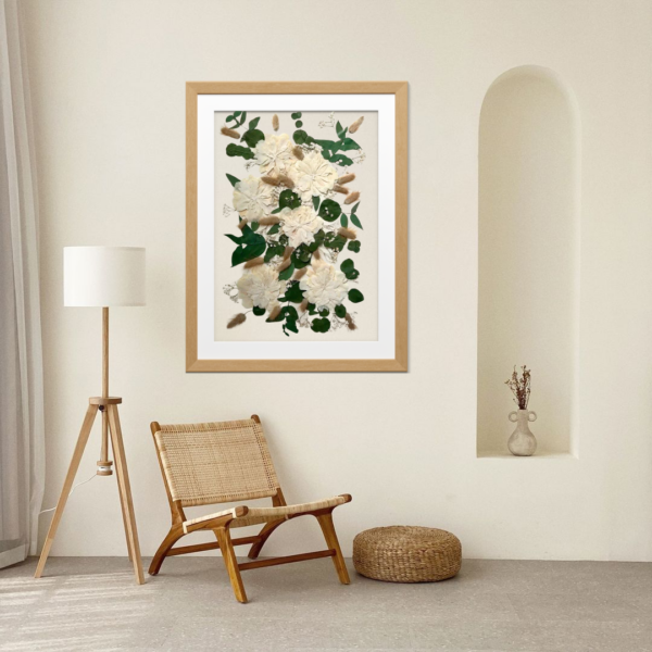 Kunstvoll gestaltetes Erinnerungsbild mit gepressten weißen Blüten, grünen Eukalyptusblättern und beigen Gräsern aus einem Brautstrauß, präsentiert in einem hellen Holzrahmen mit weißem Passepartout. Das Bild hängt an einer cremefarbenen Wand in einem minimalistisch eingerichteten Wohnraum mit Rattansessel, Dreibein-Stehlampe und rundem Sitzpouf im natürlichen Boho-Stil.
