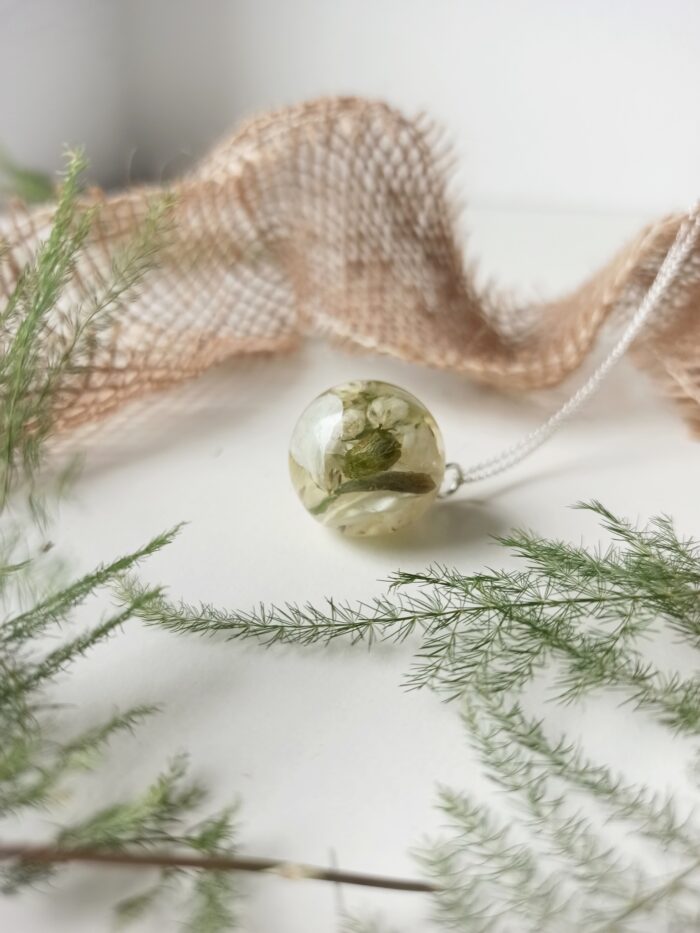 Eine minimalistische silberne Halskette mit einem transparenten Kunstharzanhänger, der zarte weiße Blüten und grüne Blätter vom Brautstrauß enthält. Die runde Harzkugel liegt auf einer weißen Oberfläche, umgeben von feinen Farnzweigen und dekorativem Juteband im oberen Bildbereich. Die schlichte, natürliche Komposition betont den botanischen Charakter des Schmuckstücks mit seinem hellen, luftigen Design. Diese Art der Brautstraußkonservierung ermöglicht es, die Frische und natürliche Schönheit der Hochzeitsblumen auf dezente Weise als tragbaren Schmuck zu bewahren.