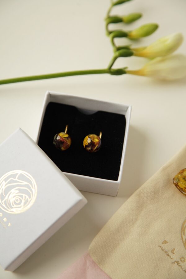 Elegante goldene Ohrringe mit kugelförmigen Kunstharzanhängern, die getrocknete Blüten in Gelb- und Rottönen enthalten. Die Ohrringe liegen in einer weißen Geschenkbox mit schwarzem Inlay. Neben der Box ist der abgenommene Deckel mit einem goldenen Rosenlogo zu sehen. Im oberen Bildbereich sind unscharfe weiße Blütenknospen an einem grünen Stängel erkennbar, während am unteren Bildrand ein beigefarbener Stoffbeutel mit einer Aufschrift zu erkennen ist. Die Aufnahme präsentiert die Ohrringe als edles Geschenk mit hochwertiger Verpackung.