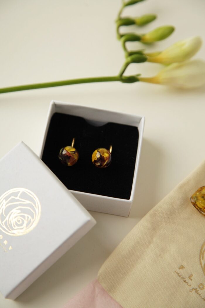 Elegante goldene Ohrringe mit kugelförmigen Kunstharzanhängern, die getrocknete Blüten in Gelb- und Rottönen enthalten. Die Ohrringe liegen in einer weißen Geschenkbox mit schwarzem Inlay. Neben der Box ist der abgenommene Deckel mit einem goldenen Rosenlogo zu sehen. Im oberen Bildbereich sind unscharfe weiße Blütenknospen an einem grünen Stängel erkennbar, während am unteren Bildrand ein beigefarbener Stoffbeutel mit einer Aufschrift zu erkennen ist. Die Aufnahme präsentiert die Ohrringe als edles Geschenk mit hochwertiger Verpackung.