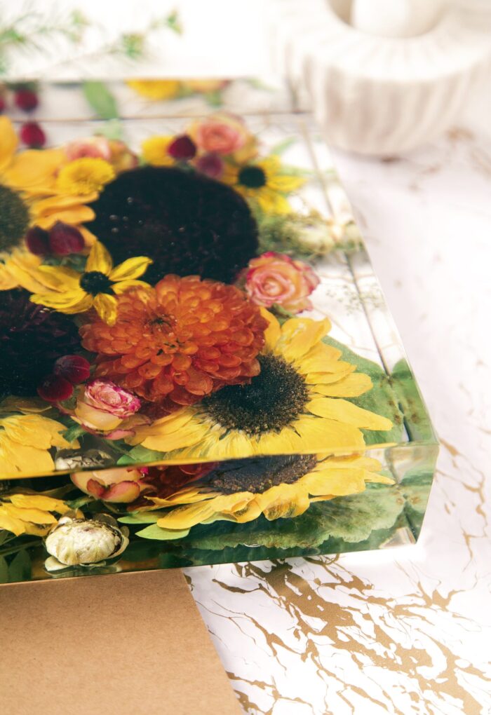 Eine Nahaufnahme eines transparenten Kunstharzblocks mit einem farbenreichen Arrangement getrockneter Herbstblumen. Der Block enthält leuchtend gelbe Sonnenblumen, eine orangefarbene Dahlie, kleine rosa Rosenblüten, dunkelrote Akzente und tiefschwarze Sonnenblumenkerne, alle in kristallklarem Harz konserviert. Die verschiedenen Blüten sind in mehreren Schichten angeordnet, was einen dreidimensionalen Tiefeneffekt erzeugt. Der Kunstharzblock ruht auf einer weißen Oberfläche mit goldenen Marmormustern, neben einem Stück braunem Kraftpapier. Im unscharfen Hintergrund ist ein weißes Keramikobjekt zu erkennen. Diese Form der Blumenkonservierung bewahrt die warmen Herbstfarben der Blüten und schafft ein dekoratives Erinnerungsstück mit leuchtenden Gelb-, Orange- und Rottönen.