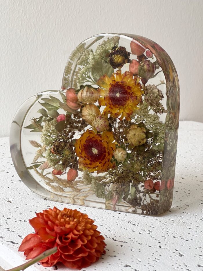 Herzförmiger Kunstharz-Block mit konservierten Trockenblumen aus einem Brautstrauß, gefüllt mit orange-gelben Strohblumen, zarten Gräsern, rosa Hagebutten und grünen Eukalyptusblättern, auf weißer Vintage-Holzoberfläche mit frischer orangefarbener Dahlienblüte im Vordergrund, natürliches Hochzeitsandenken in herbstlichen Farbtönen