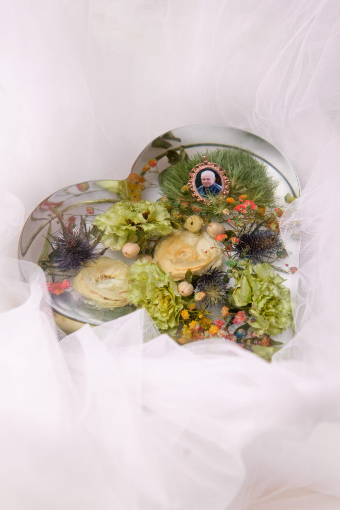 Herzförmiger Kunstharz-Anhänger mit konservierten Hochzeitsblumen aus Brautstrauß, cremefarbenen Rosen, zartgrünen Nelken und blauen Disteln, mit eingearbeitetem goldenen Medaillon-Fotorahmen für besondere Erinnerungen, auf weißem Tüllstoff arrangiert als emotionales Hochzeitsandenken