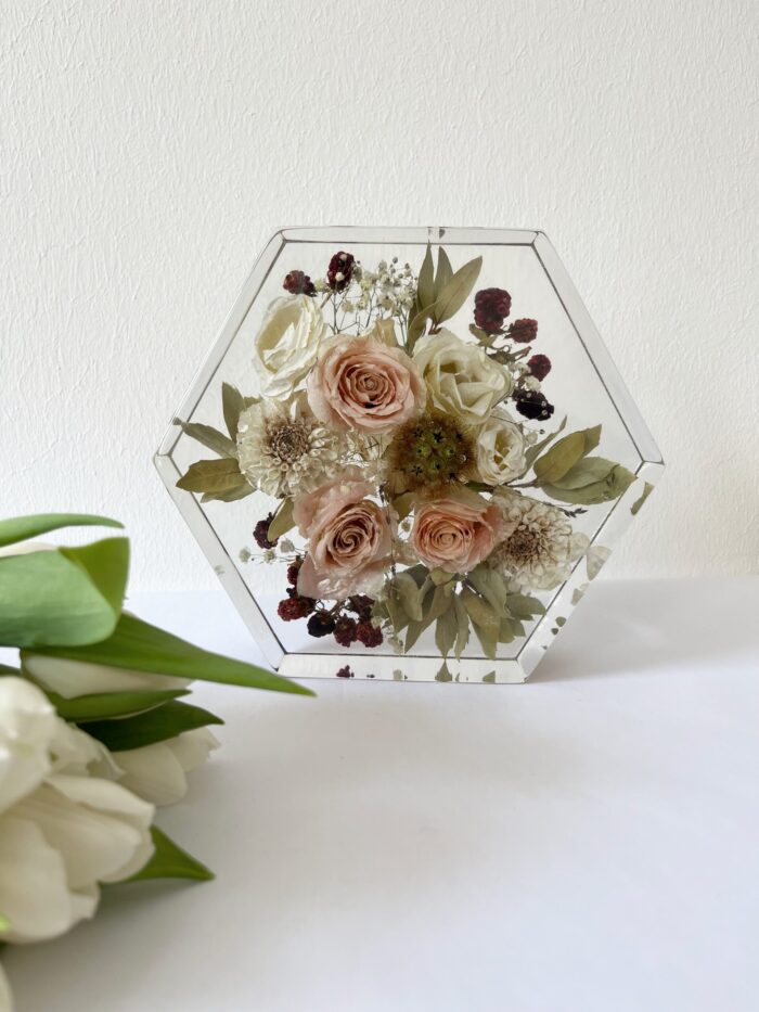 Moderner sechseckiger Kunstharz-Block mit konservierten Brautstraußblumen, gefüllt mit pudrigen rosa Rosen, weißen Blüten, kleinen burgunderroten Beeren und Eukalyptusblättern, auf weißem Tisch platziert mit frischen weißen Tulpen im Vordergrund, elegantes geometrisches Hochzeitsandenken im Boho-Vintage-Stil