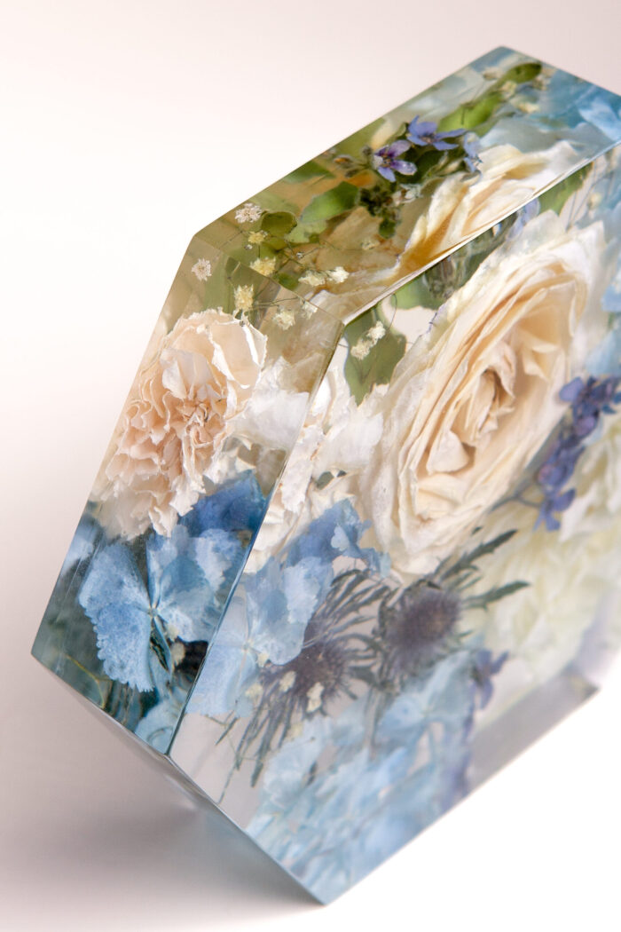 Nahaufnahme eines sechseckigen Kunstharz-Blocks mit konservierten Brautstraußblumen, detaillierte Seitenansicht mit cremefarbenen Rosen, zarten Nelken, blauen Hortensien und kleinen Schleierkrautblüten, transparentes facettiertes Design zeigt die Tiefenwirkung der eingegossenen Blüten, elegantes Hochzeitsandenken mit "Something Blue"-Farbelementen