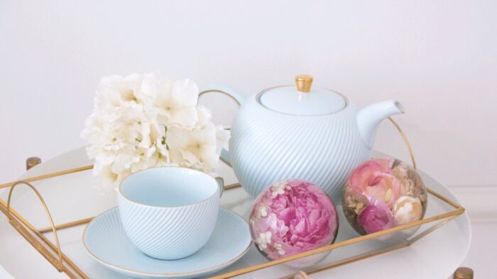 Ein elegantes Arrangement auf einem goldgerahmten Glastablett mit hellblauem Teeservice und Kunstharzkugeln mit konservierten Blumen. Das Teeservice besteht aus einer hellblauen Teekanne mit feinen diagonalen Rillen und goldenem Knopf sowie einer passenden Teetasse mit Untertasse. Daneben liegen zwei transparente Kunstharzkugeln – eine mit einer leuchtend pinken Pfingstrose und weißen Blüten, die andere mit zarten rosa und cremefarbenen Rosen. Im linken Bereich des Tabletts steht ein kleiner Strauß weißer Hortensienblüten. Das gesamte Arrangement ruht auf einer weißen Oberfläche vor einem hellen Hintergrund und verkörpert einen eleganten, femininen Einrichtungsstil, bei dem die konservierten Blumen in den Harzkugeln die Farbakzente des ansonsten pastellfarbenen Ensembles bilden.