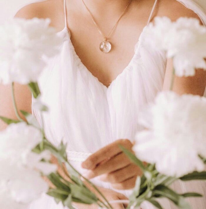 Zarter Anhänger mit konservierten weißen Hochzeitsblumen an einer goldenen Kette, getragen über einem cremefarbenen Kleid, umgeben von frischen weißen Blüten