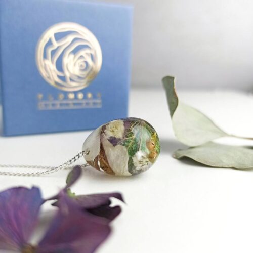 Handgefertigter Anhänger aus Epoxidharz mit eingebetteten weißen, grünen und lila Blüten an einer Silberkette, platziert vor einer blauen Geschenkbox mit goldenem Rosenlogo und getrockneten Blumenblättern