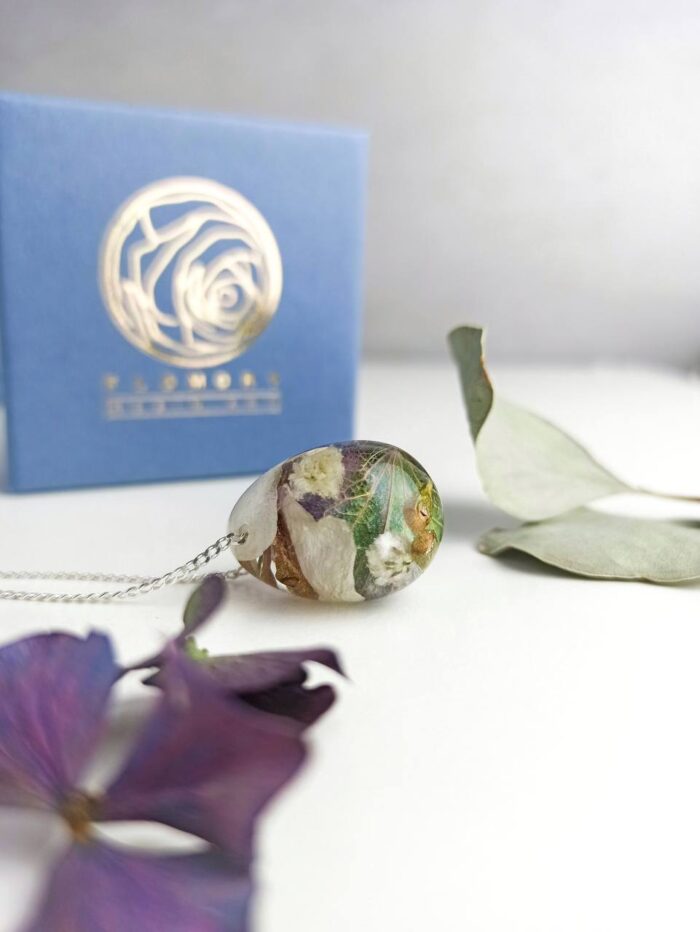 Handgefertigter Anhänger aus Epoxidharz mit eingebetteten weißen, grünen und lila Blüten an einer Silberkette, platziert vor einer blauen Geschenkbox mit goldenem Rosenlogo und getrockneten Blumenblättern