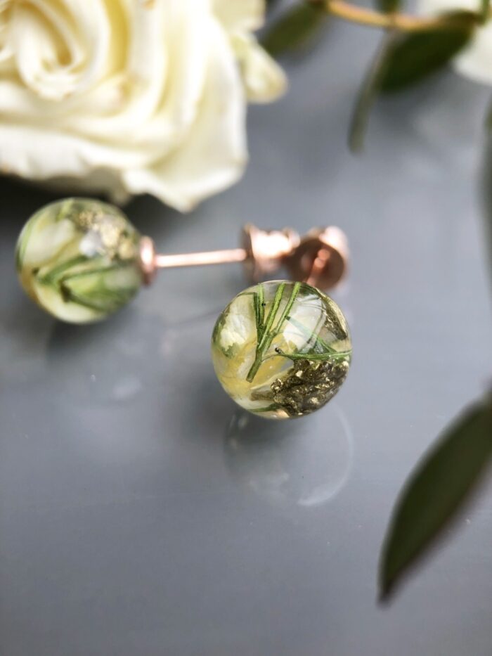 Handgefertigte Ohrstecker aus Epoxidharz mit zarten weißen und grünen Blüten, 0,8 cm rund, auf reflektierender Oberfläche mit verschwommenem grünem Hintergrund