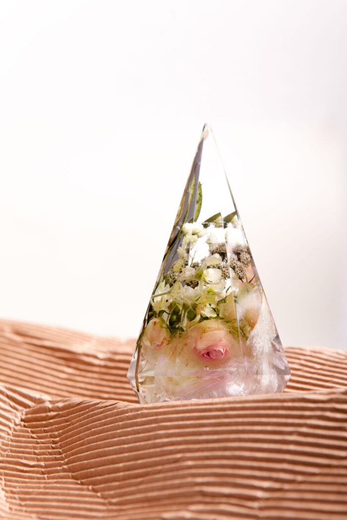 Transparente Kristallpyramide mit konservierten Brautstraußblumen auf natürlicher Holzoberfläche, eingegossene Rosen und Schleierkraut in Kunstharz, handgefertigtes Hochzeitsandenken als Tischdekoration