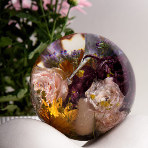Eine große transparente Kunstharzkugel mit einem farbenfrohen Arrangement getrockneter Blumen auf einer weißen geschwungenen Oberfläche. Die Kugel enthält konservierte rosa und cremefarbene Rosen, dunkelviolette Blüten und leuchtend gelbe Blumenelemente, die von zarten Schleierkrautblüten umgeben sind. Im Hintergrund steht ein Topf mit frischen rosa Chrysanthemen, deren Farbe harmonisch mit den konservierten Blüten in der Harzkugel korrespondiert. Das Licht erzeugt leichte Reflexionen auf der glatten Oberfläche der Kugel und betont die dreidimensionale Anordnung der eingebetteten Blumen. Diese Art der Blumenkonservierung dient als dekoratives Element und dauerhaftes Erinnerungsstück.