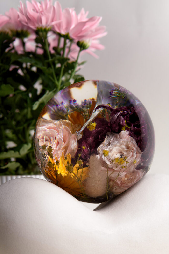 Eine große transparente Kunstharzkugel mit einem farbenfrohen Arrangement getrockneter Blumen auf einer weißen geschwungenen Oberfläche. Die Kugel enthält konservierte rosa und cremefarbene Rosen, dunkelviolette Blüten und leuchtend gelbe Blumenelemente, die von zarten Schleierkrautblüten umgeben sind. Im Hintergrund steht ein Topf mit frischen rosa Chrysanthemen, deren Farbe harmonisch mit den konservierten Blüten in der Harzkugel korrespondiert. Das Licht erzeugt leichte Reflexionen auf der glatten Oberfläche der Kugel und betont die dreidimensionale Anordnung der eingebetteten Blumen. Diese Art der Blumenkonservierung dient als dekoratives Element und dauerhaftes Erinnerungsstück.