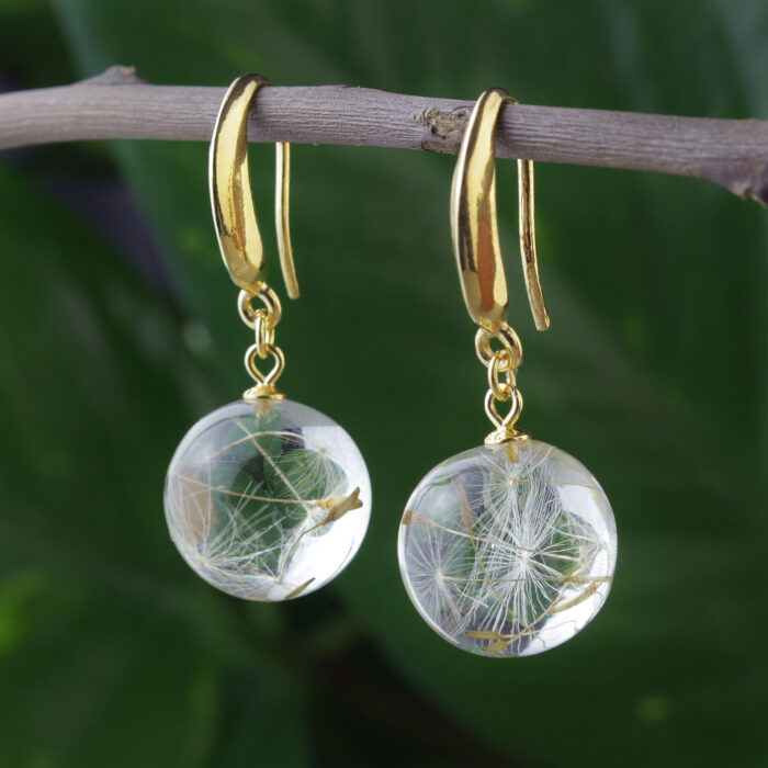 Elegante Ohrringe mit transparenten Kunstharzkugeln, in denen zarte Pusteblumensamen konserviert wurden. Die runden Kugeln hängen an goldenen Bügeln und sind auf einem natürlichen Holzzweig vor unscharfem grünem Hintergrund drapiert. Die feinen, weißen Schirmchen der Pusteblumensamen sind deutlich sichtbar im klaren Harz eingeschlossen und verleihen den Ohrringen eine luftige, naturinspirierte Ästhetik. Diese Art von Schmuck kombiniert natürliche Elemente mit moderner Konservierungstechnik und ist ein beliebtes Accessoire für Naturliebhaber oder als besonderes Geschenk.