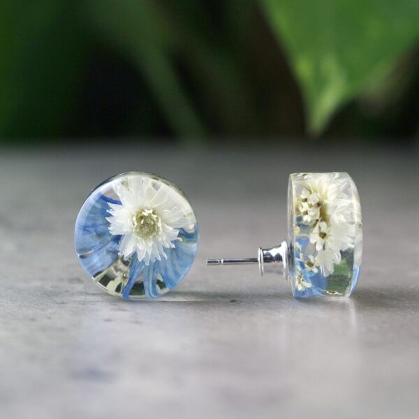 Handgefertigte Ohrstecker aus Epoxidharz mit weißer Blüte und blauen Akzenten, rund, auf grauem Untergrund mit unscharfem grünem Hintergrund, silberne Verschlüsse