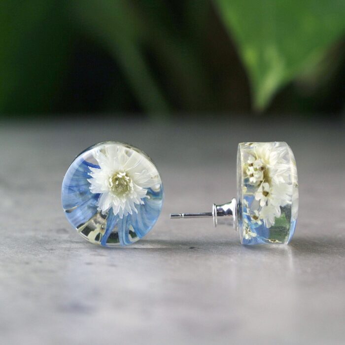 Handgefertigte Ohrstecker aus Epoxidharz mit weißer Blüte und blauen Akzenten, rund, auf grauem Untergrund mit unscharfem grünem Hintergrund, silberne Verschlüsse