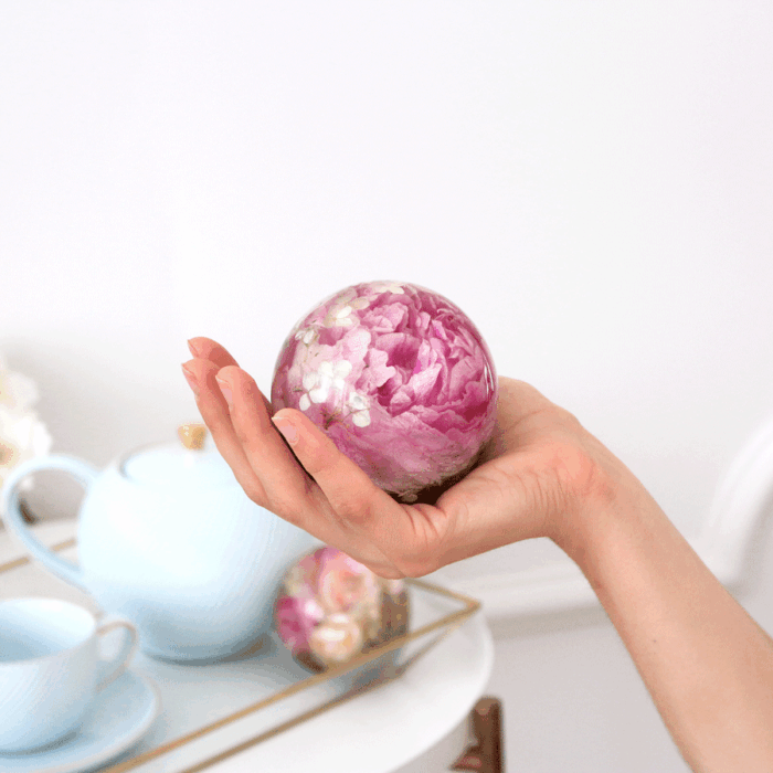 Eine Hand hält behutsam eine transparente Kunstharzkugel, in der eine vollständige pinke Pfingstrosenblüte konserviert wurde. Die Kugel zeigt die üppigen, mehrschichtigen Blütenblätter der Pfingstrose in verschiedenen Rosa- und Fuchsiatönen, die durch das klare Harz besonders lebendig wirken. Im unscharfen Hintergrund sind weiße Teetassen auf einem Tablett zu erkennen, was eine elegante, helle Wohnatmosphäre andeutet. Die Größenverhältnisse der Kugel zur Hand verdeutlichen deren handliche Dimension. Diese Art der Blumenkonservierung bewahrt die Schönheit besonderer Blüten dauerhaft und dient als dekoratives Element oder persönliches Erinnerungsstück für besondere Anlässe.