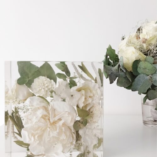 Ein rechteckiger transparenter Kunstharzblock mit konservierten weißen Blumen und grünen Eukalyptusblättern auf einer weißen Oberfläche. Der durchsichtige Block enthält cremeweiße Pfingstrosen oder Rosen, kleine weiße Blüten und verschiedene grüne Blätter, die in mehreren Schichten eingebettet sind. Rechts im Bild steht eine kleine Glasvase mit frischen weißen Rosen und Eukalyptuszweigen, die mit dem Arrangement im Harzblock harmonieren. Diese eckige Form der Blumenkonservierung bietet eine Alternative zu den häufigeren Kugelformen und eignet sich besonders gut als Tischdekoration oder Buchstütze. Der minimalistische, monochrome Farbstil in Weiß und Grün verleiht dem Arrangement eine zeitlose, elegante Ästhetik.