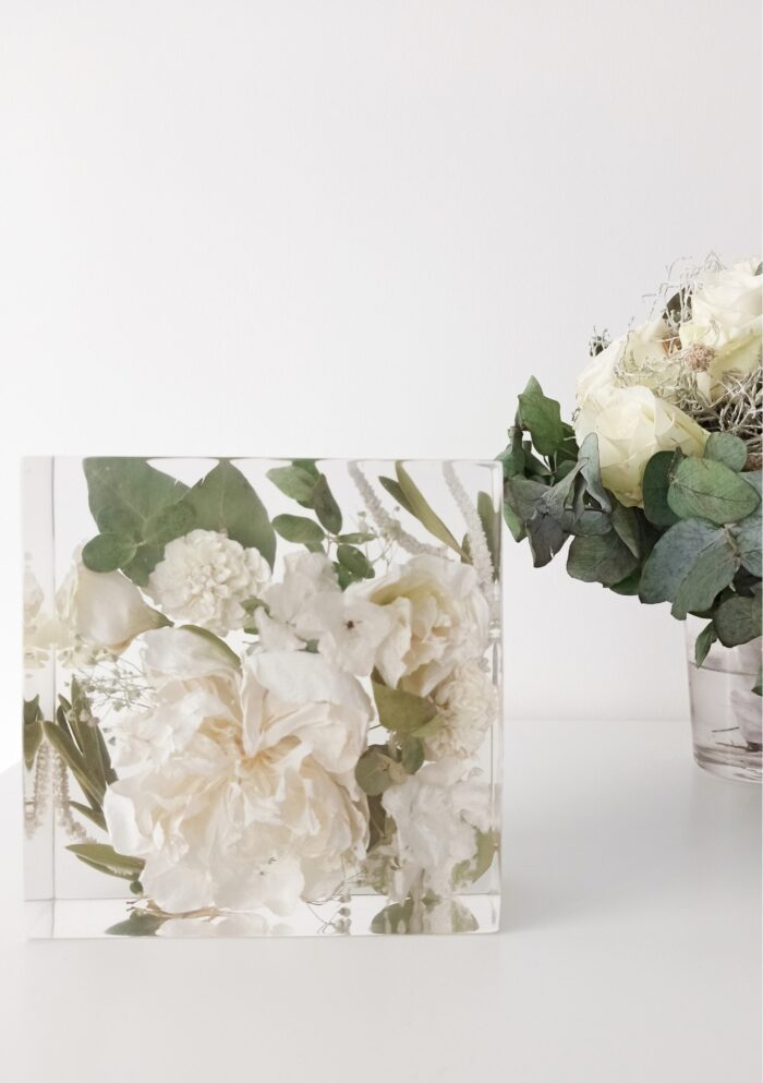 Ein rechteckiger transparenter Kunstharzblock mit konservierten weißen Blumen und grünen Eukalyptusblättern auf einer weißen Oberfläche. Der durchsichtige Block enthält cremeweiße Pfingstrosen oder Rosen, kleine weiße Blüten und verschiedene grüne Blätter, die in mehreren Schichten eingebettet sind. Rechts im Bild steht eine kleine Glasvase mit frischen weißen Rosen und Eukalyptuszweigen, die mit dem Arrangement im Harzblock harmonieren. Diese eckige Form der Blumenkonservierung bietet eine Alternative zu den häufigeren Kugelformen und eignet sich besonders gut als Tischdekoration oder Buchstütze. Der minimalistische, monochrome Farbstil in Weiß und Grün verleiht dem Arrangement eine zeitlose, elegante Ästhetik.