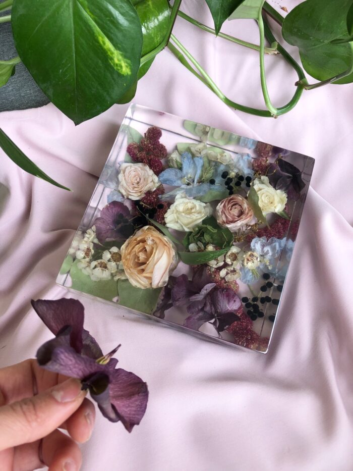 Ein quadratischer transparenter Kunstharzblock mit einem konservierten, farbenfrohen Blumenarrangement auf einem zarten rosa Stoffhintergrund. Der Block enthält apricotfarbene und cremeweiße Rosen, violette Blüten, hellblaue Blumenelemente, burgunderrote Akzente und verschiedene grüne Blätter, die in klarem Harz eingebettet sind. Im unteren Bildrand hält eine Hand eine frische violette Blüte, die mit den konservierten Blumen im Block farblich harmoniert. Im oberen Bildbereich sind große grüne Blätter einer Zimmerpflanze zu sehen. Diese eckige Form der Blumenkonservierung präsentiert das komplexe Blumenarrangement wie in einer durchsichtigen Vitrine und bewahrt die natürliche Schönheit und Farbvielfalt des ursprünglichen Straußes. Der Block eignet sich als dekoratives Element oder als Erinnerungsstück an besondere Anlässe