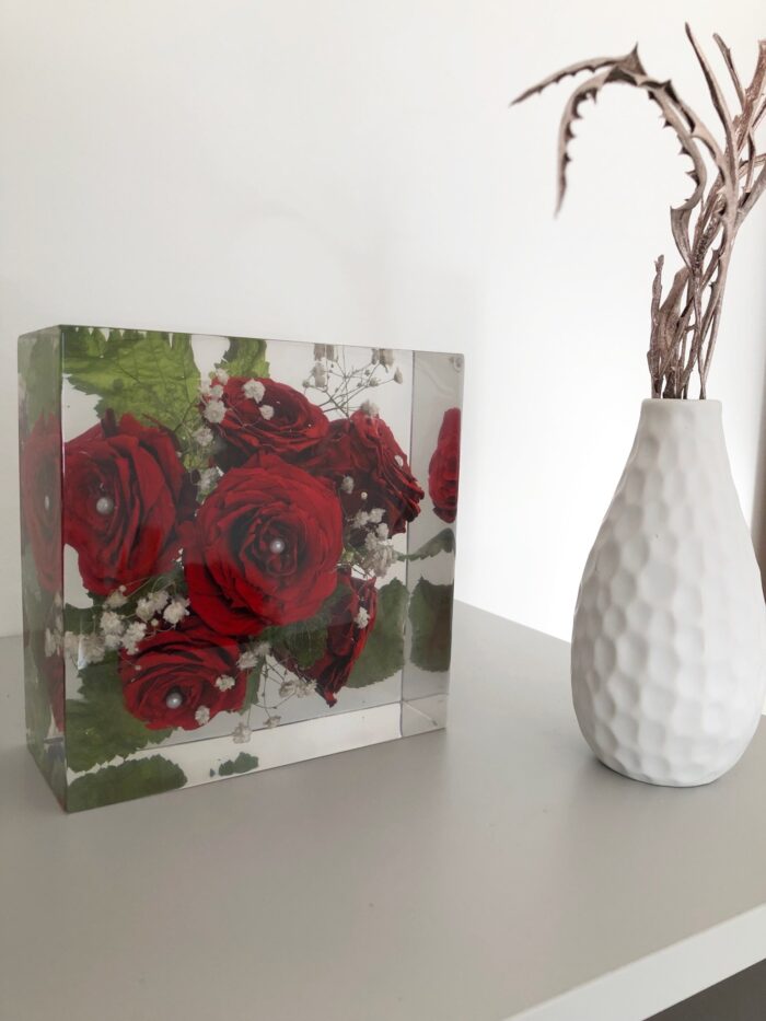 Ein quadratischer transparenter Kunstharzblock mit konservierten leuchtend roten Rosen, Schleierkraut und grünen Blättern, auf einer weißen Oberfläche platziert. Die tiefroten Rosen sind vollständig im klaren Harz eingebettet und behielten ihre intensive Farbe und Form. Rechts daneben steht eine moderne, weiße Keramikvase mit strukturierter Oberfläche, in der getrocknete Zweige arrangiert sind. Die Komposition zeigt den Kontrast zwischen den dauerhaft konservierten Blumen im Kunstharzblock und der minimalistischen, modernen Vasengestaltung. Der Block dient als dekoratives Erinnerungsstück, das möglicherweise einen besonderen Anlass wie einen Hochzeitstag oder Jahrestag symbolisiert. Das Arrangement steht vor einer schlichten weißen Wand und verkörpert einen zeitgemäßen, eleganten Einrichtungsstil.