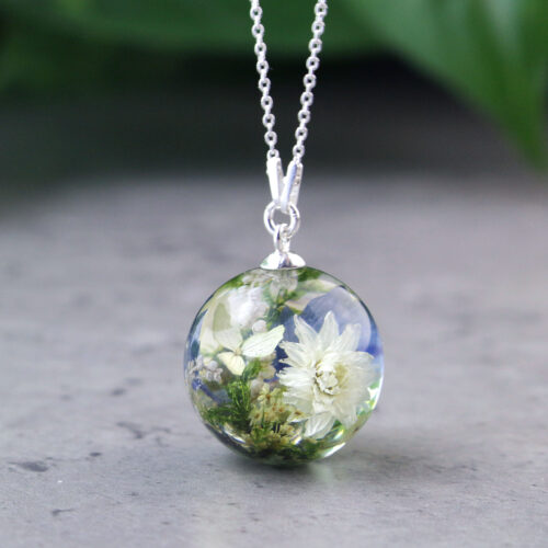 Eine silberne Halskette mit einem runden Kunstharzanhänger, der Blüten aus einem Brautstrauß konserviert. In der transparenten Kugel sind eine weiße Chrysantheme oder Dahlie, kleine weiße Blüten wie Schleierkraut und zarte grüne Blätter zu sehen, die perfekt im klaren Harz erhalten bleiben. Der Anhänger hängt an einer feinen silbernen Gliederkette und liegt auf einer grauen, strukturierten Oberfläche. Im unscharfen Hintergrund ist ein grüner Farbton erkennbar. Dieser handgefertigte Schmuck ermöglicht es Bräuten, die Blumen ihres Hochzeitstages als persönliches Erinnerungsstück dauerhaft zu bewahren und zu tragen.