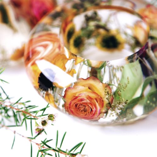 Handgefertigter Teelichthalter aus transparentem Kunstharz in Armreif-Form, gefüllt mit konservierten orangegelben Rosen und Sonnenblumen aus einem Brautstrauß, umgeben von frischen Kräutern und Grün, romantische Hochzeitserinnerung als stimmungsvolle Tischdekoration