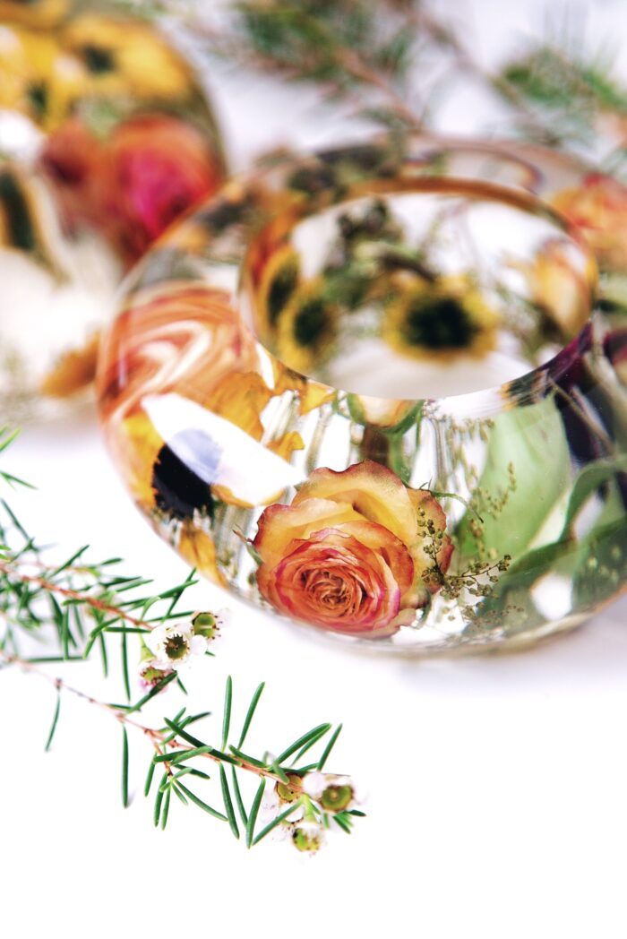 Handgefertigter Teelichthalter aus transparentem Kunstharz in Armreif-Form, gefüllt mit konservierten orangegelben Rosen und Sonnenblumen aus einem Brautstrauß, umgeben von frischen Kräutern und Grün, romantische Hochzeitserinnerung als stimmungsvolle Tischdekoration