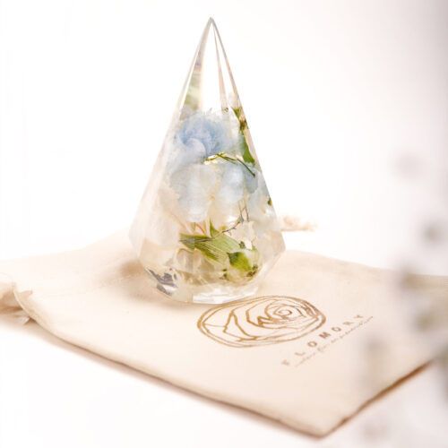 Tropfenförmiger Kunstharz-Kristall mit konservierten weißen und hellblauen Hortensien aus Brautstrauß, präsentiert auf elegantem Stoffbeutel mit goldgeprägtem Rosenlogo, exklusives Hochzeitsandenken als Geschenk verpackt