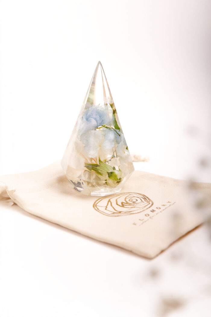 Tropfenförmiger Kunstharz-Kristall mit konservierten weißen und hellblauen Hortensien aus Brautstrauß, präsentiert auf elegantem Stoffbeutel mit goldgeprägtem Rosenlogo, exklusives Hochzeitsandenken als Geschenk verpackt