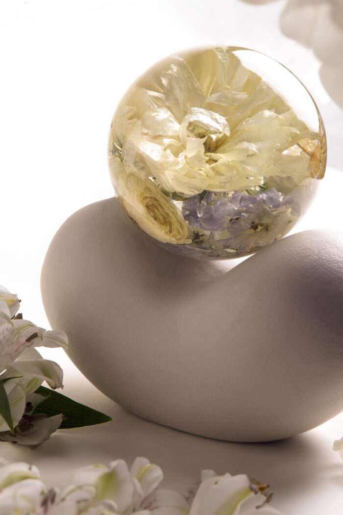 Eine transparente Kunstharzkugel mit konservierten cremefarbenen Blüten und zarten violetten Akzenten, platziert auf einem modernen, geschwungenen matten Keramikständer in Weiß. Die Kugel enthält mehrere eingebettete Blüten, darunter weiße Rosen und kleine lila Blütenelemente, die in verschiedenen Schichten im Harz konserviert wurden. Im unteren Bildbereich sind frische weiße Lilienblüten zu sehen, die eine natürliche Ergänzung zur konservierten Blumenkomposition bilden. Das weiche Licht erzeugt subtile Schatten und hebt die transparente Qualität der Harzkugel hervor, während der minimalistische Keramikständer einen eleganten Kontrast zur natürlichen Blütenform bietet. Diese dekorative Kombination verkörpert einen zeitgemäßen, reduzierten Stil für die dauerhafte Präsentation konservierter Blumen.