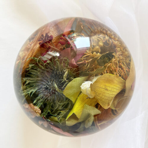 Eine transparente Kunstharzkugel gefüllt mit verschiedenen getrockneten Wildblumen und botanischen Elementen in warmen Herbstfarben. Im Zentrum ist eine dunkle Distel oder ähnliche stachelige Blüte zu sehen, umgeben von gelben Blütenblättern, burgunderroten Blütenelementen und verschiedenen getrockneten Gräsern und Kräutern in Goldtönen. Die Kugel liegt auf einem weichen, weißen Stoffhintergrund, der leichte Falten wirft. Die Lichtreflexionen auf der Harzkugel betonen die strukturellen Details der konservierten Pflanzen im Inneren. Diese Art der Blumenkonservierung bewahrt die natürliche Textur und Farbe der Wildblumen in einem dauerhaften Medium und schafft ein rustikales, naturnahes Dekorationselement.