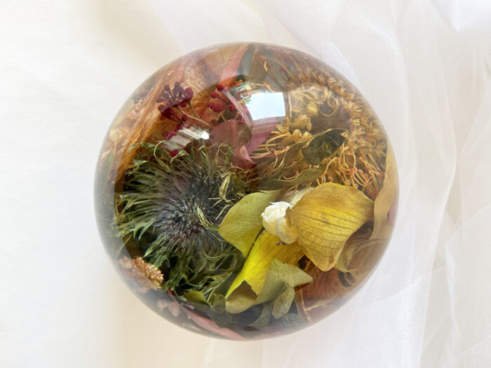 Eine transparente Kunstharzkugel gefüllt mit verschiedenen getrockneten Wildblumen und botanischen Elementen in warmen Herbstfarben. Im Zentrum ist eine dunkle Distel oder ähnliche stachelige Blüte zu sehen, umgeben von gelben Blütenblättern, burgunderroten Blütenelementen und verschiedenen getrockneten Gräsern und Kräutern in Goldtönen. Die Kugel liegt auf einem weichen, weißen Stoffhintergrund, der leichte Falten wirft. Die Lichtreflexionen auf der Harzkugel betonen die strukturellen Details der konservierten Pflanzen im Inneren. Diese Art der Blumenkonservierung bewahrt die natürliche Textur und Farbe der Wildblumen in einem dauerhaften Medium und schafft ein rustikales, naturnahes Dekorationselement.